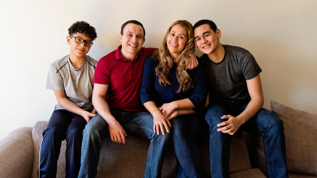 Family photography consisting of 4 family members sat on the sofa and smiling for a photograph. A young boy is on the left, with his dad next to him. The mother is next to the dad, and lastly, on the far right is another teenage boy, older than the first. 