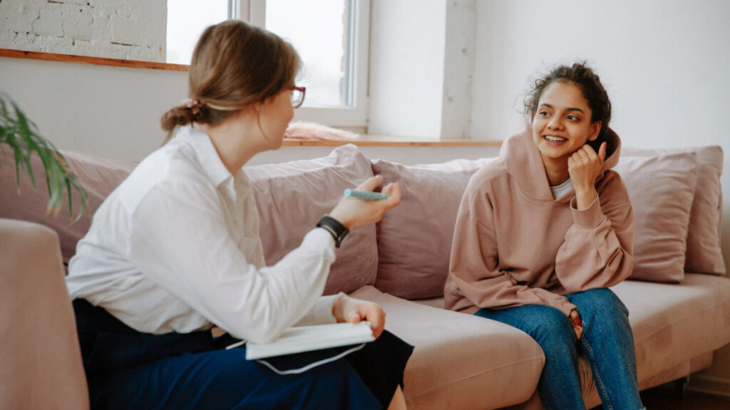 Image of 2 people talking whilst sat on opposite ends of the sofa. One lady is wearing a white shirt and is holding notepad and pen. The other is a teenage girl who is wearing a pink hoodie and jeans, smiling at the woman. 