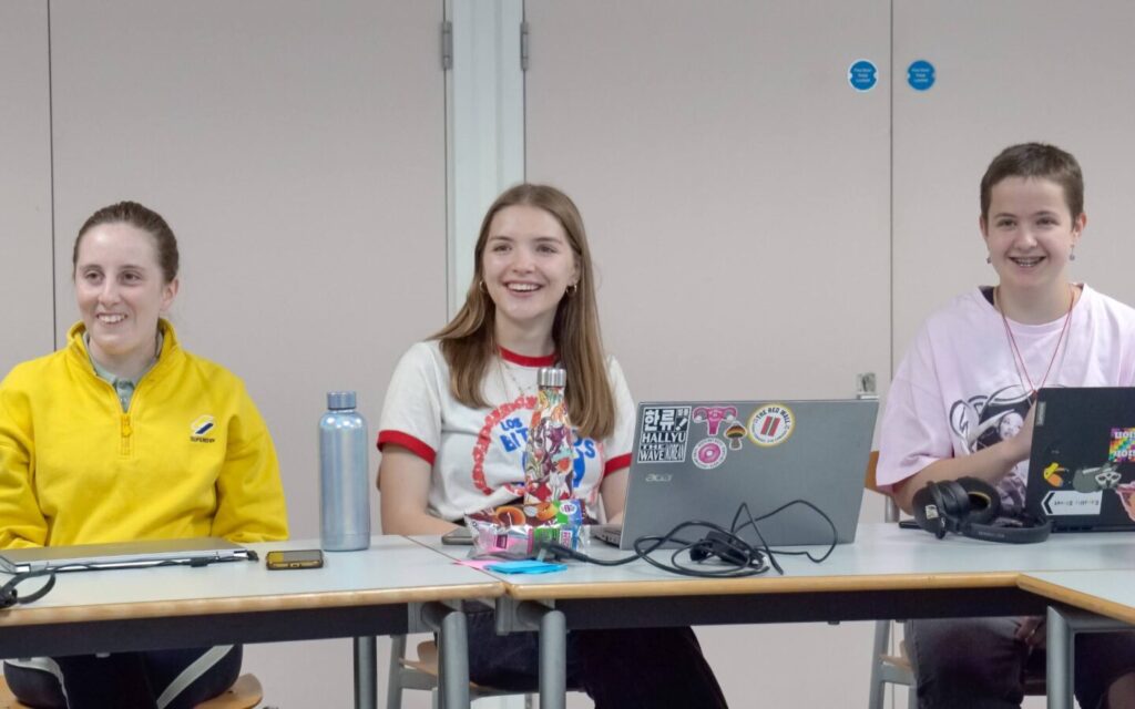 Three young people sat behind a desk smiling, two behind open laptops